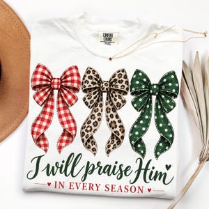 May include: A white t-shirt featuring three large bows in red gingham, leopard print, and green with white polka dots. Below the bows, the text reads, "I will praise Him IN EVERY SEASON" in dark green and red script and block lettering.