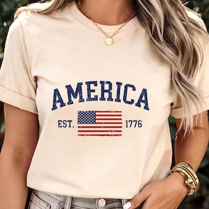 May include: A cream-coloured short-sleeve t-shirt featuring the word AMERICA in distressed navy blue lettering above a graphic of the United States flag. The text EST. 1776 is printed in navy blue on either side of the flag graphic on the chest.