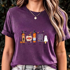 May include: A heather purple short-sleeve t-shirt featuring a graphic of five anthropomorphic school supplies dressed for Halloween: a crayon, a mummy apple, a glue stick, a spiral notebook, and a pencil ghost, all wearing witch hats.