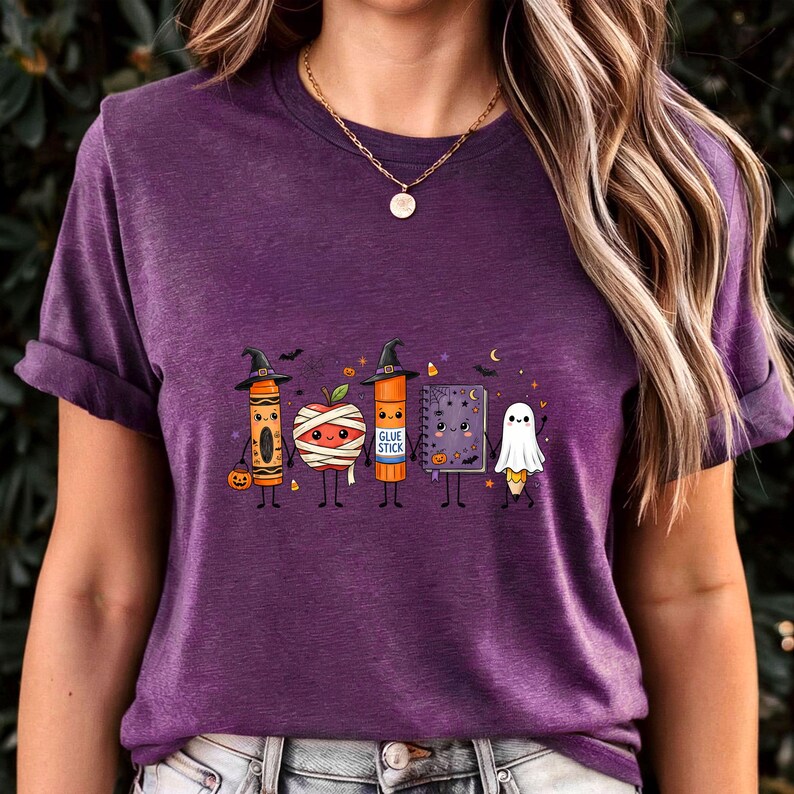 May include: A heather purple short-sleeve t-shirt featuring a graphic of five anthropomorphic school supplies dressed for Halloween: a crayon, a mummy apple, a glue stick, a spiral notebook, and a pencil ghost, all wearing witch hats.