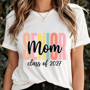 May include: A white short-sleeved t-shirt featuring the word SENIOR in large, colorful block letters behind the black cursive text Mom and the black printed text class of 2027. The design uses pastel shades of orange, yellow, teal, and pink.