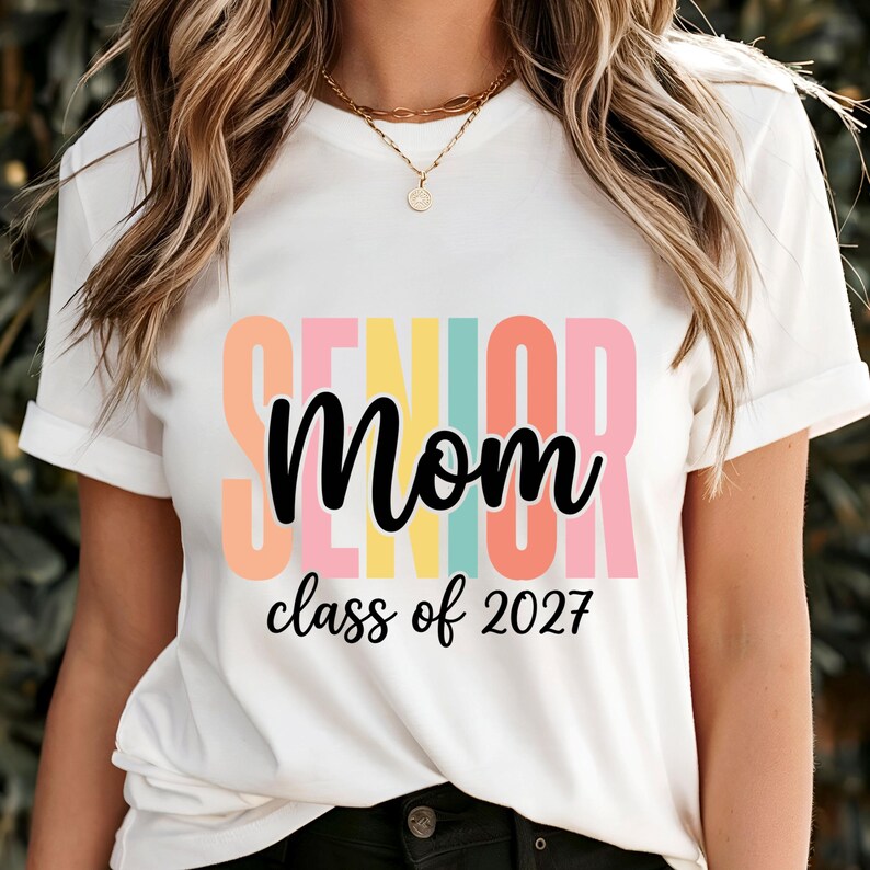 May include: A white short-sleeved t-shirt featuring the word SENIOR in large, colorful block letters behind the black cursive text Mom and the black printed text class of 2027. The design uses pastel shades of orange, yellow, teal, and pink.
