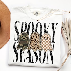 May include: A white t-shirt featuring the black distressed text SPOOKY SEASON and three ghost illustrations. The ghosts have different patterns: one with green camouflage, one with a brown leopard print, and one with a tan and white checkerboard.
