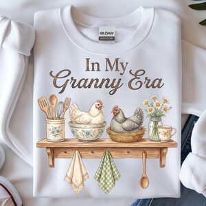 May include: White sweatshirt with the text "In My Granny Era". The graphic features a wooden shelf with a hen in a bowl, a hen in a nest, daisies, and kitchen utensils. Two decorative towels and a wooden spoon hang below.