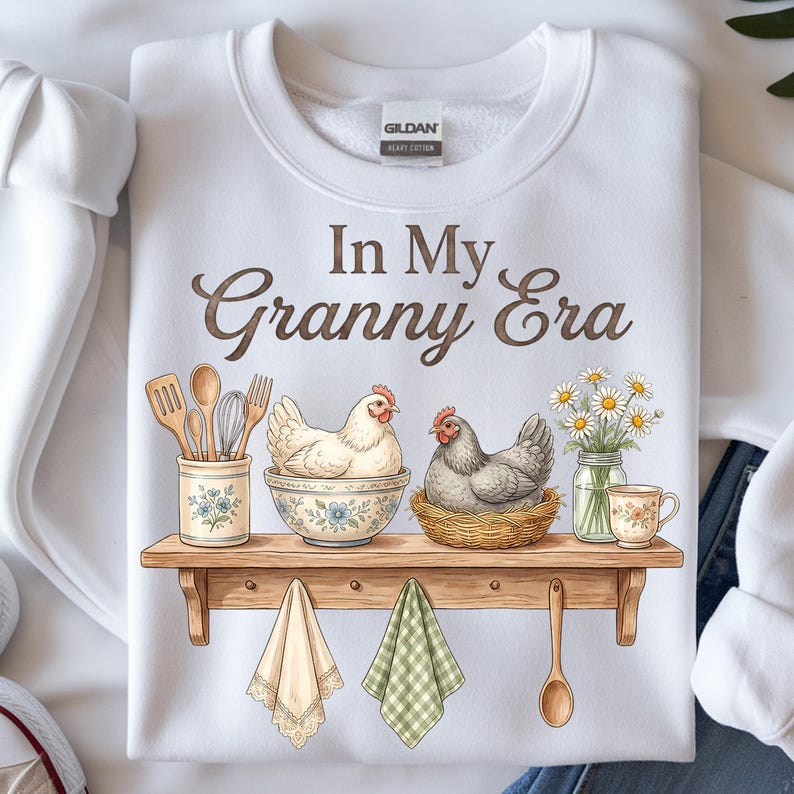 May include: White sweatshirt with the text "In My Granny Era". The graphic features a wooden shelf with a hen in a bowl, a hen in a nest, daisies, and kitchen utensils. Two decorative towels and a wooden spoon hang below.