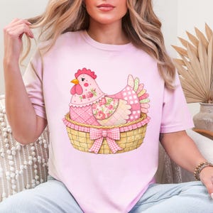 May include: A light pink t-shirt with a colourful illustration of a hen in a basket. The hen is decorated with floral and patchwork patterns in pinks, reds, and yellows. The basket has a gingham bow. The design is charming and cheerful, perfect for a casual look.