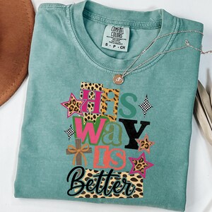 May include: A teal Comfort Colors t-shirt featuring the text "His Way Is Better" in a vibrant, patterned design. The design incorporates stars, a cross, and leopard print elements. A silver necklace with a pendant is draped across the shirt.