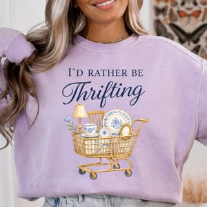 Op de afbeelding: Een lichtpaarse trui met de marineblauwe tekst "I'D RATHER BE Thrifting". De afbeelding toont een gouden winkelwagentje gevuld met vintage servies, een lamp en een kleine stoel.