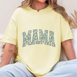 May include: A light yellow t-shirt with the word "MAMA" in a floral design across the chest. The floral design features small white flowers on a blue-green background. The shirt has short sleeves and a relaxed fit.
