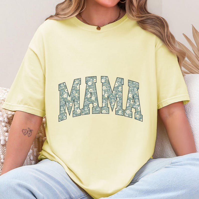 May include: A light yellow t-shirt with the word "MAMA" in a floral design across the chest. The floral design features small white flowers on a blue-green background. The shirt has short sleeves and a relaxed fit.
