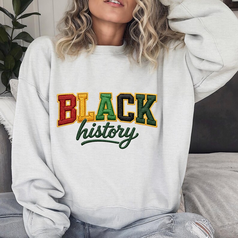 May include: A light gray sweatshirt with the words "BLACK history" in a colorful, embroidered design. The text is in a bold, collegiate-style font, with the word "BLACK" in red, gold, green, and black. The word "history" is in green script.