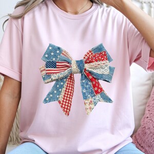 May include: A light pink t-shirt featuring a large bow design. The bow is made of various fabrics, including denim, red and white gingham, and floral patterns. The bow incorporates an American flag and lace accents.