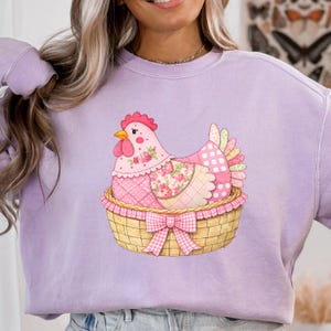 May include: A lilac sweatshirt with a charming chicken illustration. The chicken is pink, adorned with floral and polka dot patterns, nestled in a woven basket with a pink gingham bow. The design is centrally placed on the garment.