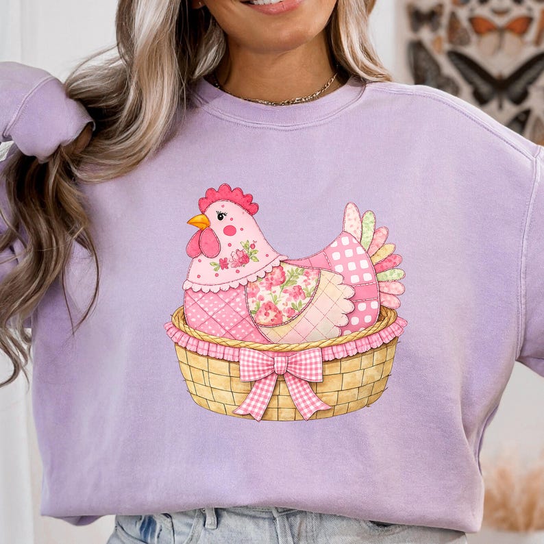 May include: A lilac sweatshirt with a charming chicken illustration. The chicken is pink, adorned with floral and polka dot patterns, nestled in a woven basket with a pink gingham bow. The design is centrally placed on the garment.