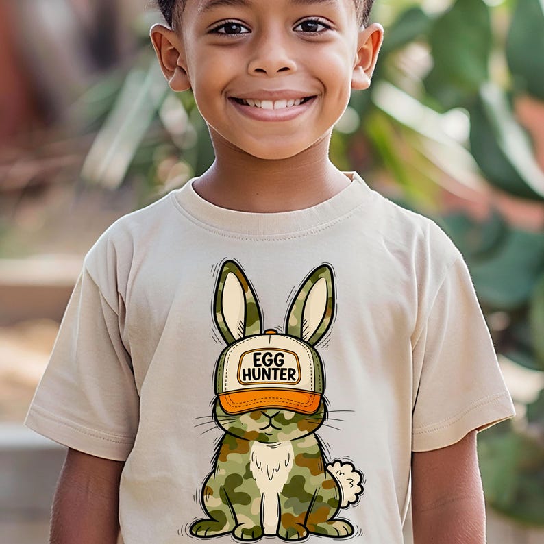Puede incluir: Una camiseta beige con un gr&aacute;fico de un conejo que lleva un sombrero con la inscripci&oacute;n "EGG HUNTER". El dise&ntilde;o del conejo incorpora un patr&oacute;n de camuflaje. La camiseta la lleva un ni&ntilde;o.