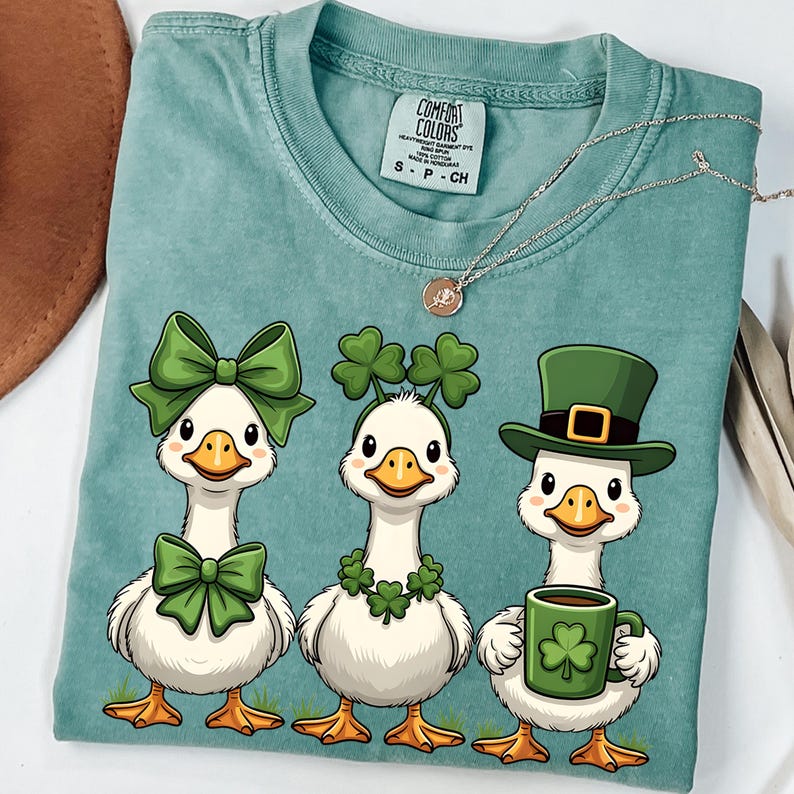 May include: A teal Comfort Colors t-shirt featuring three cartoon geese. Each goose is decorated with St. Patrick's Day themed accessories, including green bows, shamrock headbands, and a green hat. One goose holds a mug with a shamrock.