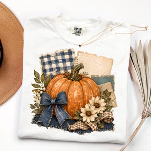 May include: A white t-shirt featuring a graphic print of a large orange pumpkin, a blue denim-textured bow, white berries, and two light-colored flowers. The design includes blue gingham and lined paper elements with torn edges on a dark denim base.