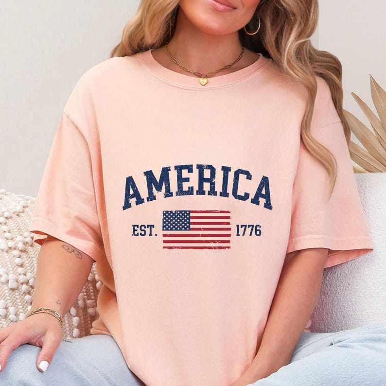 May include: A peach-coloured short-sleeve t-shirt featuring the dark blue text AMERICA arched above a graphic of the United States flag. The text EST. 1776 is printed in dark blue on either side of the flag, all with a distressed, weathered texture.