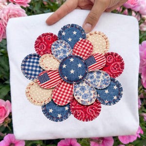 May include: White sweatshirt featuring a patriotic flower design. The flower petals are made of various fabrics, including denim, gingham, and paisley, in red, white, and blue. The center of the flower has white stars on a blue background.