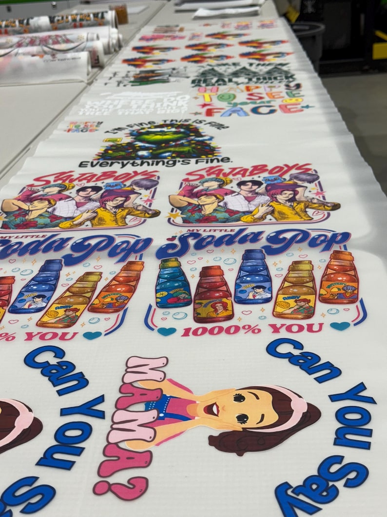May include: White printed fabric displaying multiple designs. The designs include text such as "Everything's Fine" and "My Little Soda Pop," alongside cartoon characters and soda bottle illustrations. The fabric is laid out on a long table.