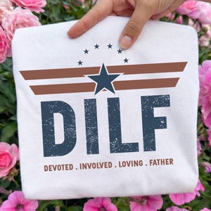May include: White t-shirt with a distressed navy blue graphic that reads "DILF" above the words "DEVOTED . INVOLVED . LOVING . FATHER." The design includes a navy blue star and brown stripes, with a circle of stars above the star.