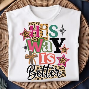 May include: White t-shirt featuring the phrase "His Way Is Better" in a vibrant, layered design. The design incorporates leopard print, stars, a cross, and a palette of colours including pink, green, and black. The shirt is presented on a woven basket.