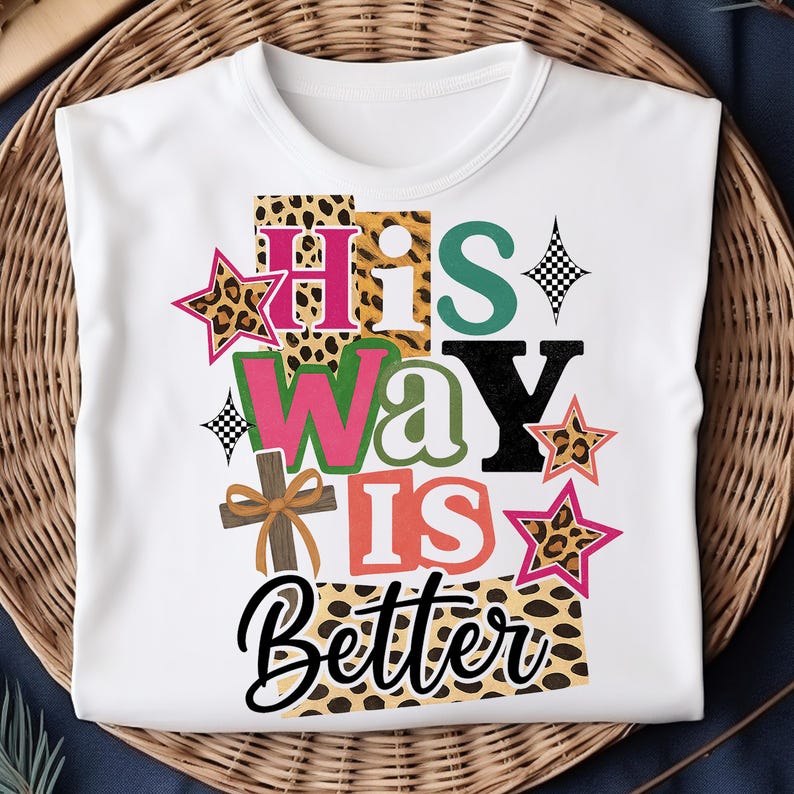 May include: White t-shirt featuring the phrase "His Way Is Better" in a vibrant, layered design. The design incorporates leopard print, stars, a cross, and a palette of colours including pink, green, and black. The shirt is presented on a woven basket.