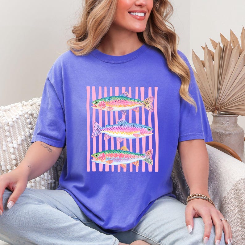 May include: A periwinkle blue t-shirt featuring a graphic of three colorful fish with pink vertical stripes in the background. The fish have rainbow hues and are arranged vertically. The shirt has short sleeves and a relaxed fit.