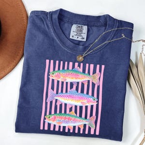 May include: Navy blue t-shirt with a graphic of three rainbow trout within a pink striped rectangle. The shirt has a crew neck and short sleeves. A gold necklace with a pendant is draped across the shirt. The brand label "Comfort Colors" is visible.