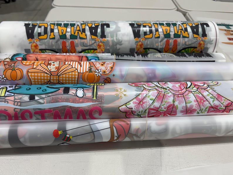 May include: A collection of rolled printed designs. One roll displays "JAMAICA" in green, yellow, and black. Other designs include pumpkins, bows, mushrooms, and floral patterns. The rolls are of varying sizes and colours.