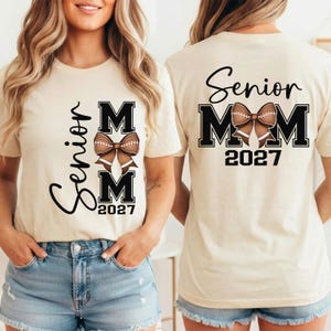 May include: A cream-colored t-shirt featuring a graphic design with the text Senior MOM 2027 and a brown football-textured bow. The front displays the text vertically, while the back shows the text horizontally with the bow centered between the M letters.