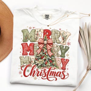 May include: A white t-shirt featuring a graphic print of the text Merry Merry Merry Christmas in a quilted pattern of red, green, and white plaid and floral fabrics. A central Christmas tree illustration is topped with a red plaid bow.
