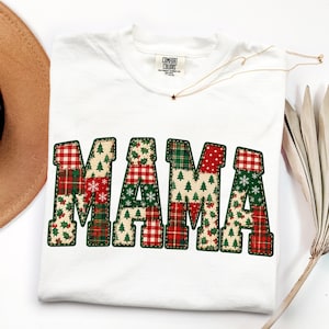 May include: A white short-sleeve t-shirt featuring the word MAMA in large block letters. The letters are filled with a patchwork pattern of red and green plaid, Christmas trees, snowflakes, and holly berries, all outlined in dark green stitching.