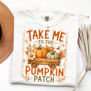 May include: A white t-shirt featuring a graphic of a wooden wagon filled with orange and green pumpkins, autumn leaves, and flowers. The text reads "TAKE ME TO THE PUMPKIN PATCH" in orange and green lettering, surrounded by small star accents.