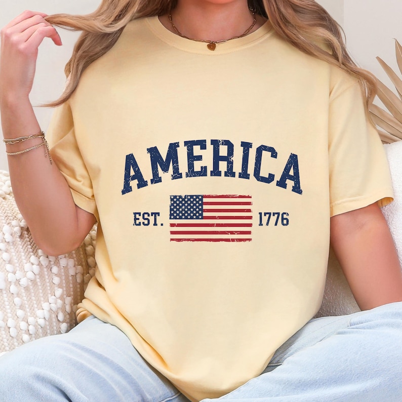 May include: A pale yellow short-sleeve t-shirt featuring the dark blue text AMERICA in a curved font above a graphic of the United States flag. The text EST. 1776 is printed on either side of the flag, all with a distressed, weathered texture.