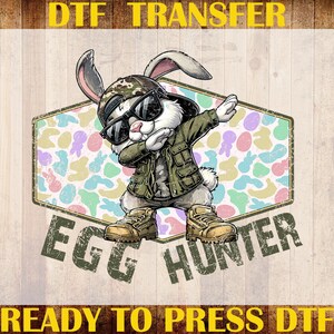 May include: A DTF transfer featuring a cartoon rabbit wearing sunglasses, a camouflage hat, and a green jacket, dabbing. The text "EGG HUNTER" is below the rabbit, and the text "DTF TRANSFER" and "READY TO PRESS DTF" are above and below the image.