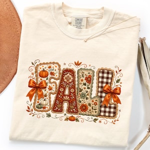 May include: A cream-colored t-shirt featuring the word FALL in large, decorative letters. Each letter has a unique pattern including pumpkins, floral motifs, and brown gingham. Two orange satin bows are attached to the F and L letters.