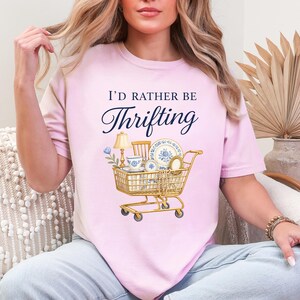 Op de afbeelding: Lichtroze T-shirt met de tekst "I'd Rather Be Thrifting" boven een illustratie van een winkelwagentje gevuld met vintage items. De kar bevat een lamp, kopjes, borden en een spiegel.