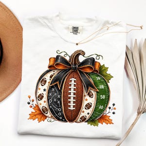 May include: A white t-shirt featuring a graphic of a pumpkin designed like a football. The pumpkin has a brown football texture, a black section with white play diagrams, and a green section with yard lines. It is accented with a black bow and leaves.