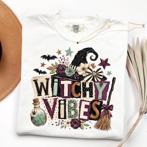 May include: A white t-shirt featuring a graphic print with the text WITCHY VIBES. The design includes a black witch hat, purple and green flowers, a potion bottle, a broom, bats, and stars in shades of purple, green, black, and tan.