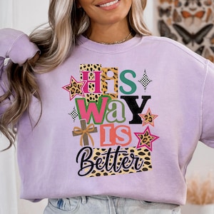 May include: A lilac sweatshirt featuring the text "His Way Is Better" in a layered design. The design incorporates leopard print, stars, and a cross.