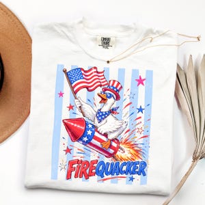 May include: A white t-shirt featuring a graphic of a duck wearing a patriotic top hat, sunglasses, and bandana, riding a firework rocket while holding an American flag. The text FIREQUACKER is printed below in red and blue letters.