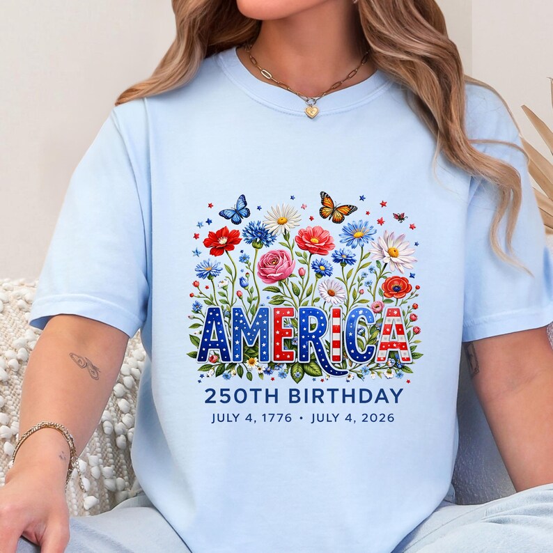 K&ouml;nnte beinhalten: Hellblaues T-Shirt mit floralem Design und dem Wort "AMERICA" in Rot, Wei&szlig; und Blau. Das Shirt feiert den 250. Geburtstag mit den Daten 4. Juli 1776 und 4. Juli 2026.