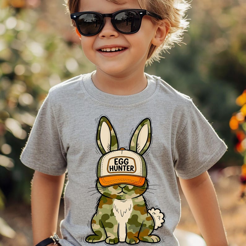 Puede incluir: Una camiseta gris claro con un conejito de dibujos animados que lleva un sombrero con el texto "EGG HUNTER". El conejito tiene un patr&oacute;n de camuflaje en tonos verdes, marrones y beige. El sombrero es naranja y blanco. La camiseta es de manga corta.