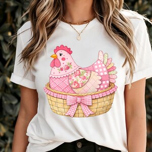 May include: White t-shirt with a colourful chicken illustration. The chicken is pink with floral and polka dot patterns, nestled in a woven basket with a pink gingham bow. The design is detailed and vibrant.