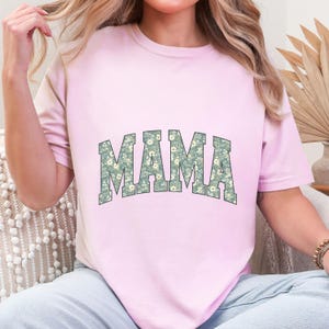 May include: A light pink t-shirt with the word "MAMA" in large, block letters across the front. The letters are filled with a floral pattern in shades of green and white. The shirt has short sleeves and a crew neck.
