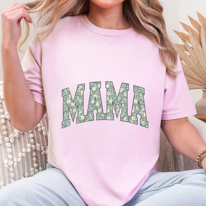 May include: A light pink t-shirt with the word "MAMA" in large, block letters across the front. The letters are filled with a floral pattern in shades of green and white. The shirt has short sleeves and a crew neck.