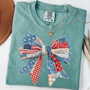 May include: A seafoam green Comfort Colors t-shirt featuring a large bow design made of patchwork denim, red gingham, floral fabric, and lace. The bow incorporates an American flag. A silver necklace with a pendant is visible.