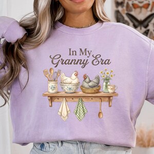 May include: A light purple sweatshirt with the text "In My Granny Era" in brown script. The graphic features a wooden shelf with kitchen utensils, chickens, flowers, and hanging towels. A wooden spoon hangs below the shelf.
