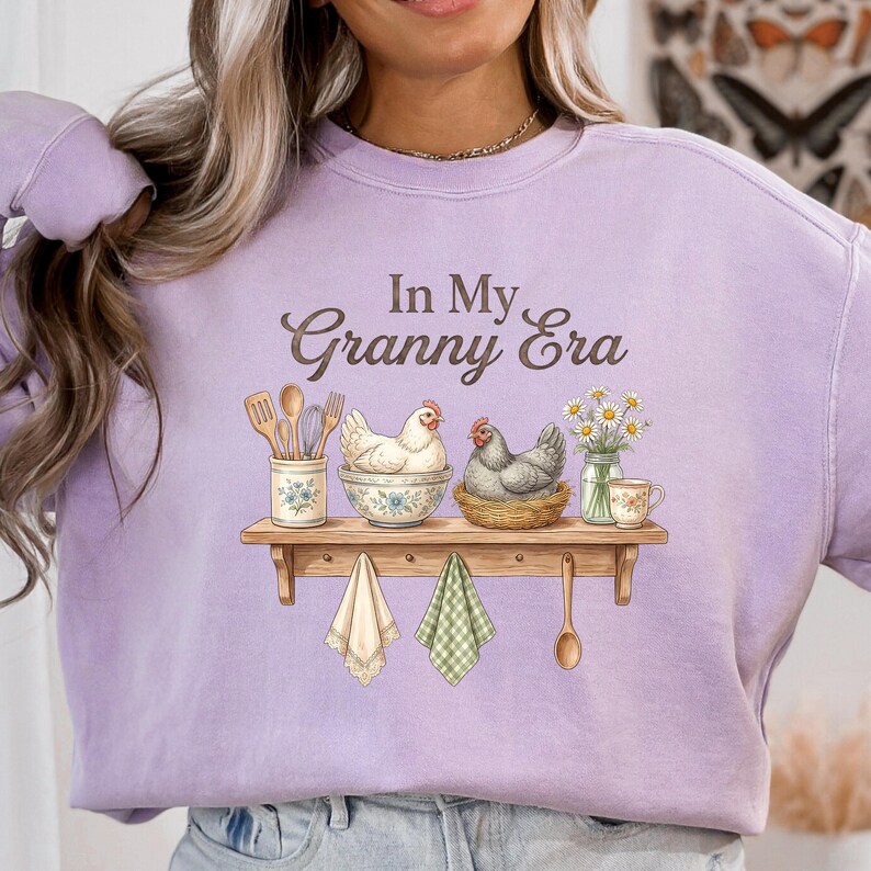 May include: A light purple sweatshirt with the text "In My Granny Era" in brown script. The graphic features a wooden shelf with kitchen utensils, chickens, flowers, and hanging towels. A wooden spoon hangs below the shelf.